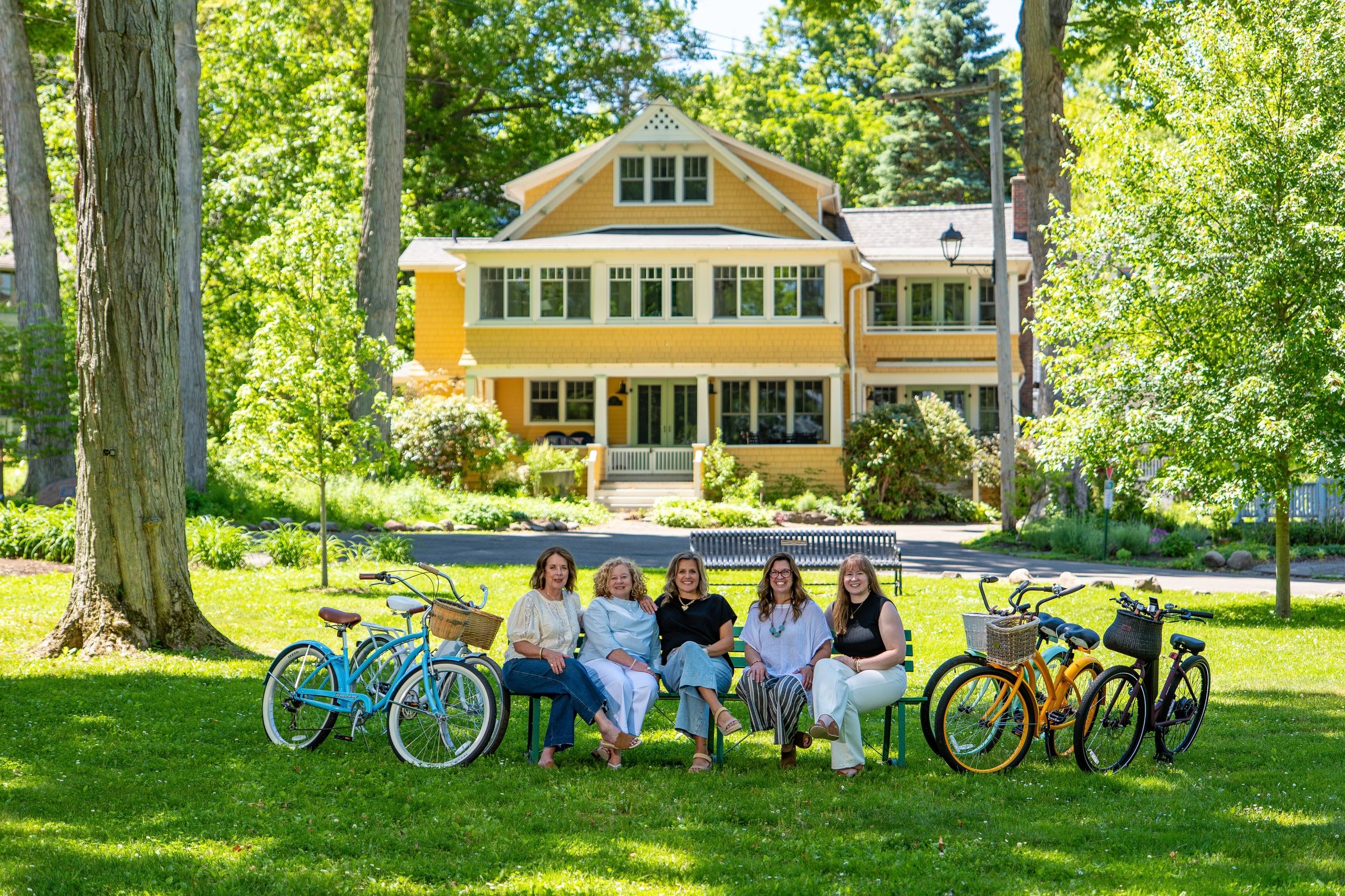 Five women sit on a bench in front of a yellow house with bikes around them.