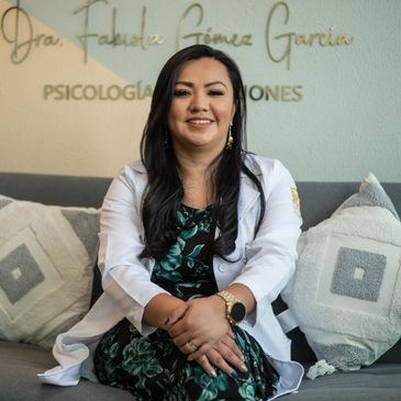 A smiling woman in a white coat sitting on a couch with decorative pillows.