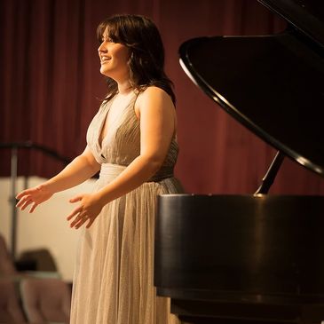 Ms. Hanna- a white woman with dark brown hair- sings while standing in a white dress. 