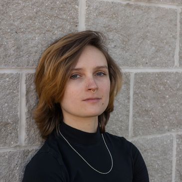 Ms. Ashley- a white woman with short brown hair- leans against a grey cinder block wall 