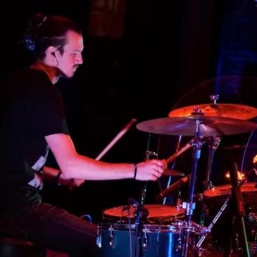 Mr. Griffin- a white man with brown hair- plays the drum set in a red lit dark room. 