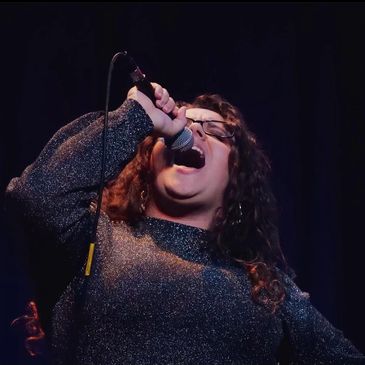 Ms. Emilierose- a white woman with long curly brown hair- sings into a mic 