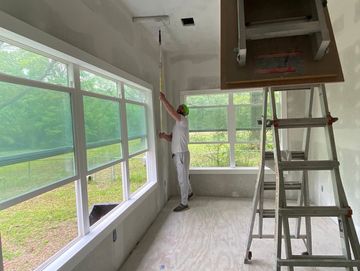 Painter applying white paint to ceiling near large windows.
