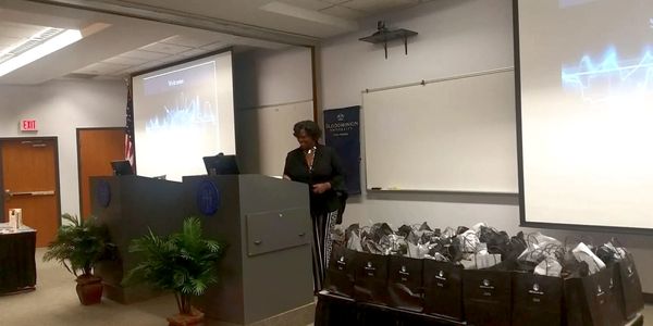 A speaker at a podium in a classroom with gift bags on a table.