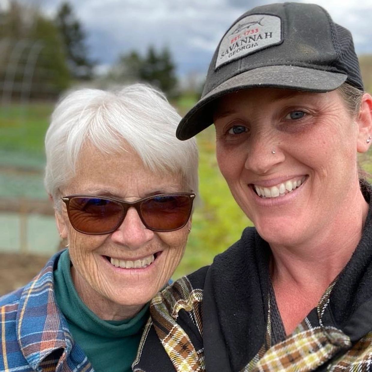 Two smiling women posing outdoors, one wearing sunglasses and the other a cap.