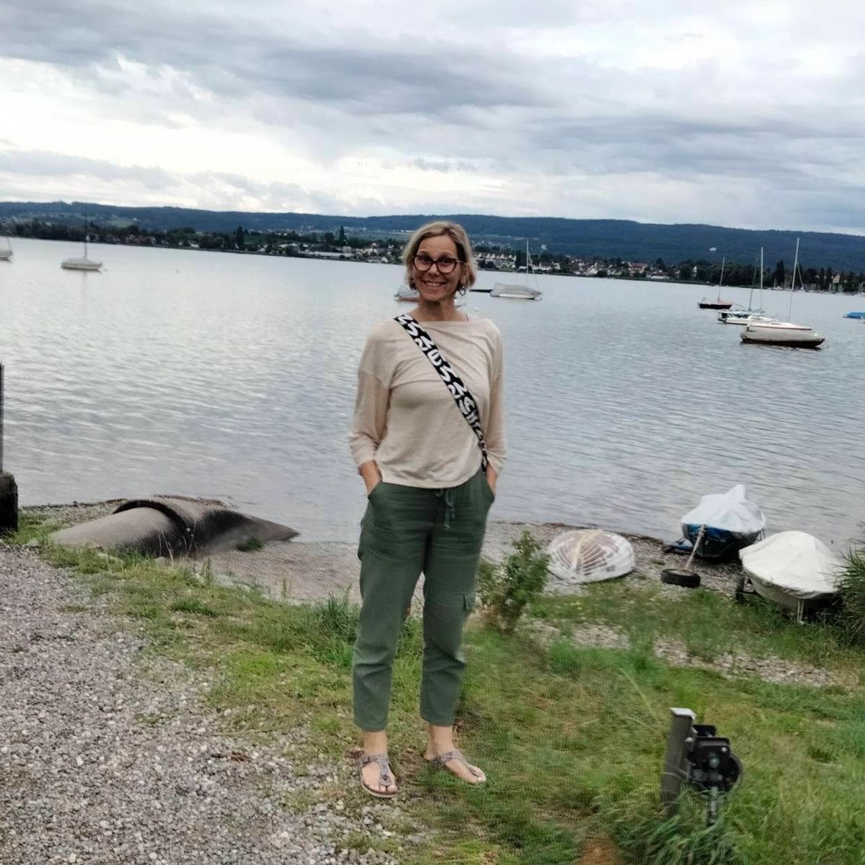 A woman stands smiling by a lakeside with boats and cloudy skies.