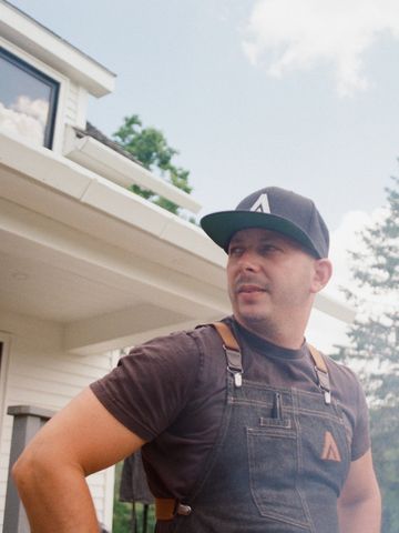 Man wearing a black cap and denim overalls standing outside a house.