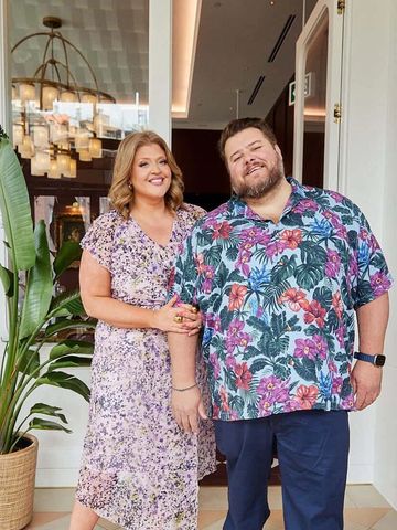 A smiling couple stands together indoors, dressed in colorful floral outfits.