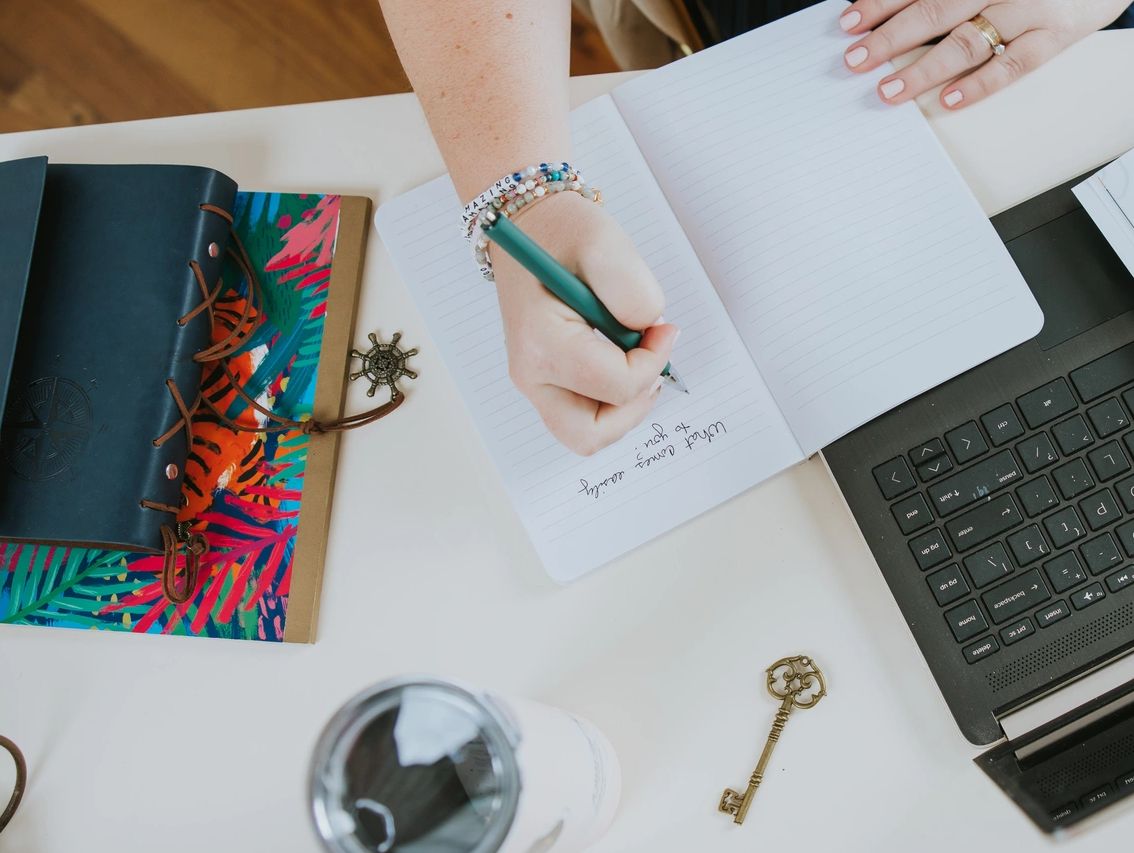 Person writing in a notebook beside a laptop on a desk.