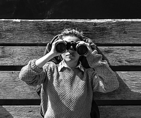 Person lying on wooden floor looking through binoculars in black and white.
