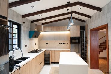 Modern kitchen with wooden cabinets and white countertops under exposed wooden beams.