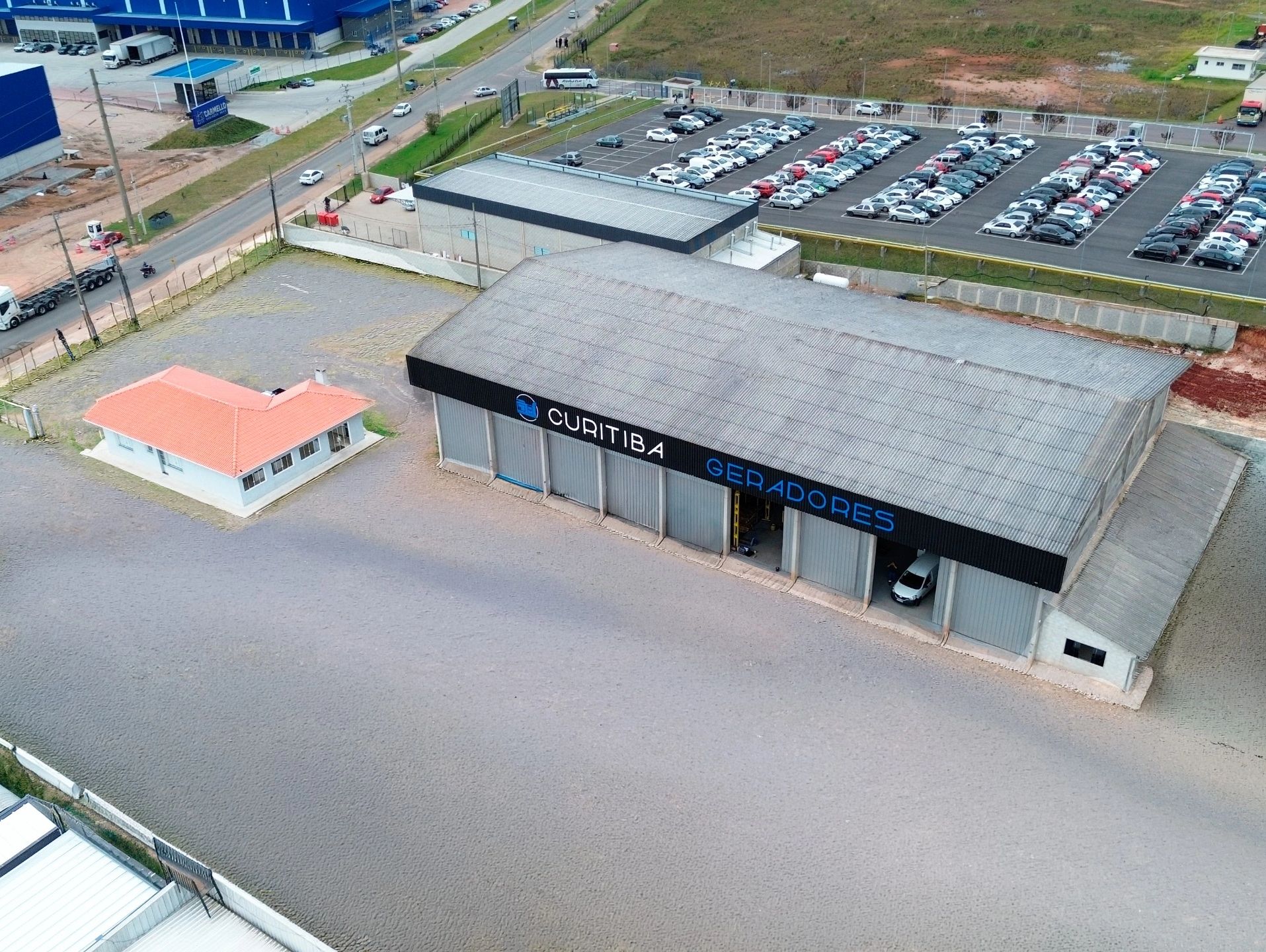 Visão aérea de uma fábrica em Curitiba com estacionamento e edifício pequeno.
