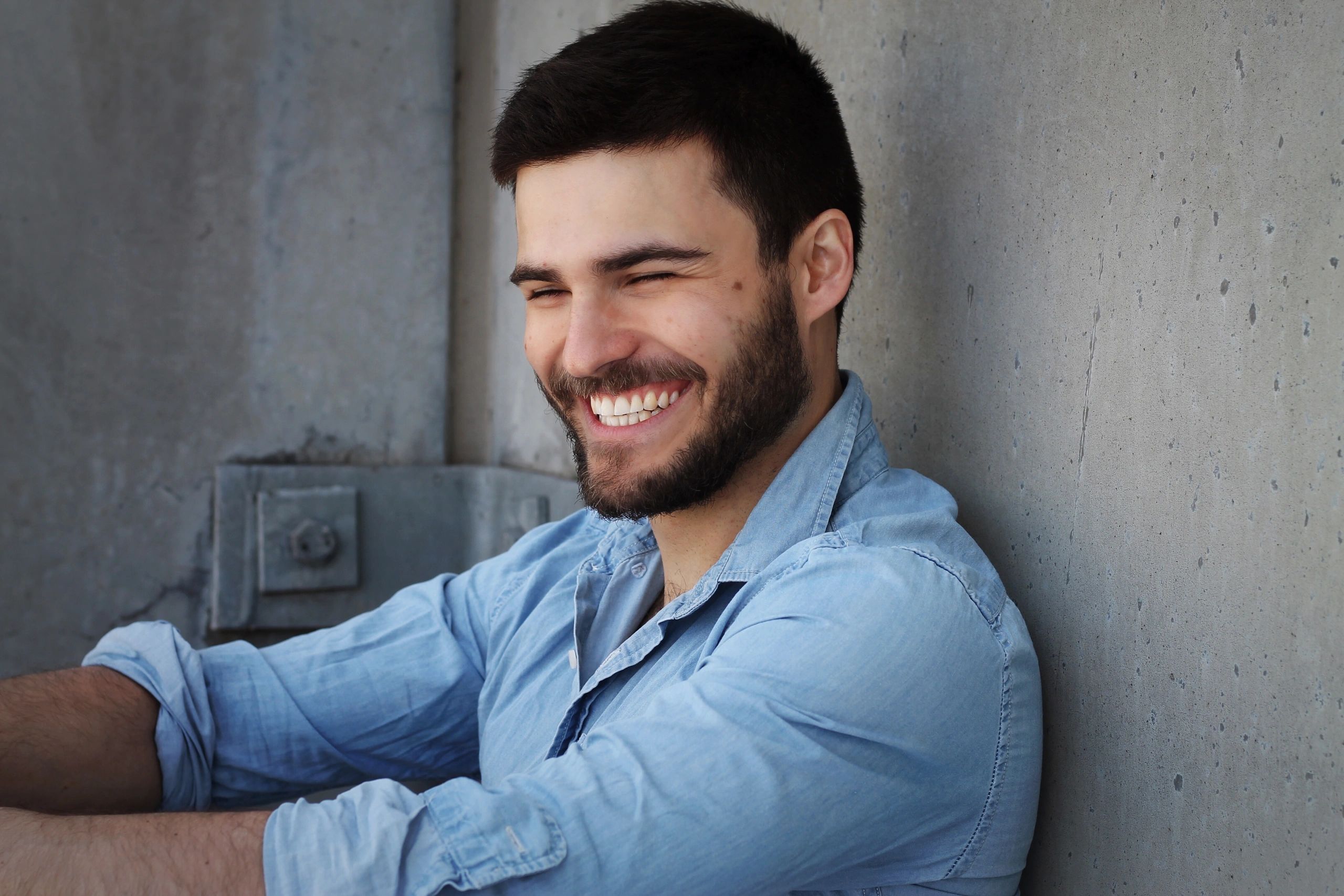 A man smiling warmly against a concrete wall.