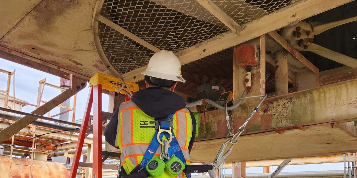 Worker in safety gear inspects industrial equipment on a ladder.