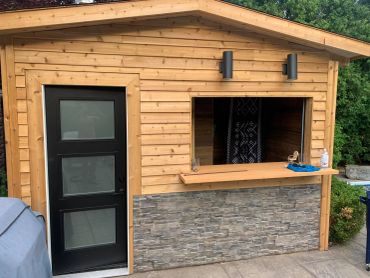 A wooden outdoor bar with a stone base and a black-framed glass door.