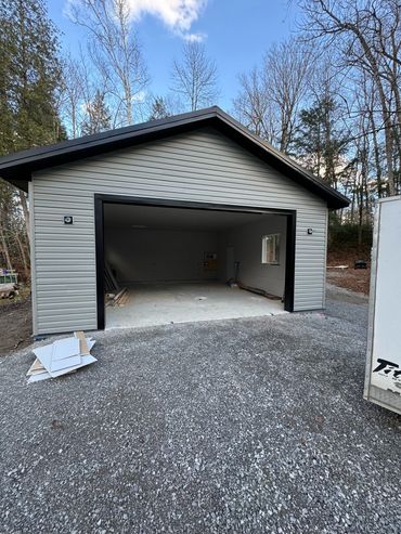Newly built single-car garage with gravel driveway, surrounded by trees under a clear sky.