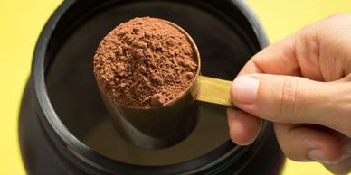 Hand holding a scoop of chocolate protein powder over a container.