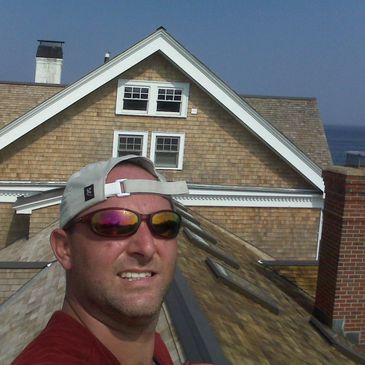 Man in sunglasses and cap taking a selfie on a rooftop with a house in the background.