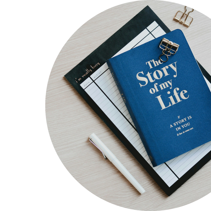 A blue book titled 'The Story of my Life' with a white pen and a planner on a light wooden table.