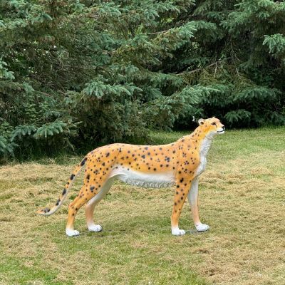 A lifelike cheetah sculpture stands on grass near pine trees.