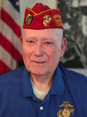 Elderly Marine Corps veteran wearing a red hat and blue shirt.