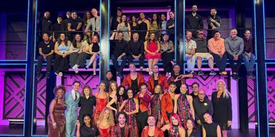A large, diverse group posing on a brightly lit stage with purple and blue lights.