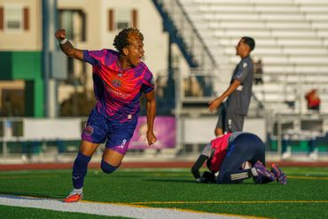Soccer player in purple and red celebrates on the field as opponents react.