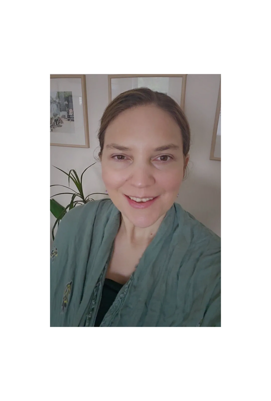 Smiling woman wearing a green shawl indoors with framed pictures and a plant behind her.