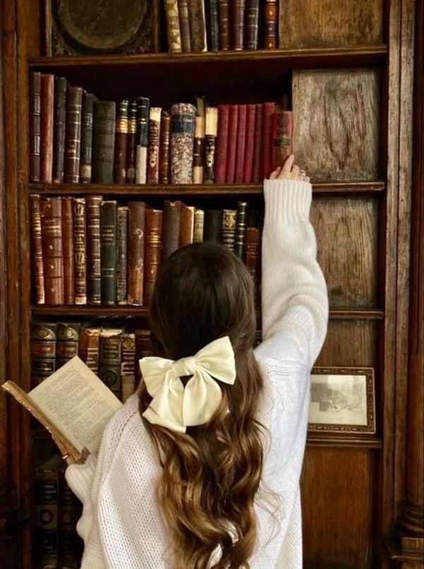Woman with curly hair and a big bow reaches for a book on a wooden bookshelf.