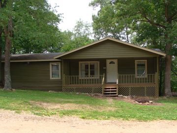Small green cabin surrounded by trees with a front porch and white door.