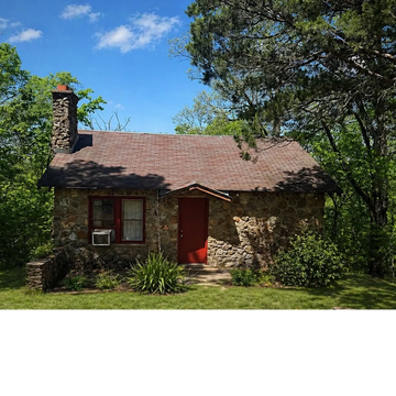 A quaint stone cottage with a red door surrounded by greenery under a clear blue sky.