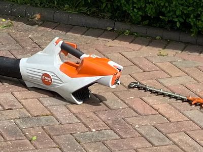 A STIHL hedge trimmer lying on a paved surface near some greenery.