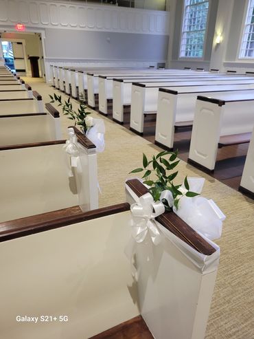 Decorated white church pews with greenery and white ribbons.