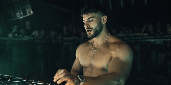 Shirtless male DJ performing at a dimly lit club.