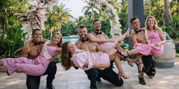 Groomsmen in bow ties holding bridesmaids in pink dresses under a floral arch.