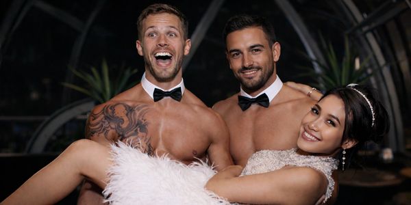 Two shirtless men in bow ties holding a smiling woman in a white dress.