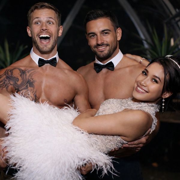 Two shirtless men in bow ties holding a smiling woman in a white dress.