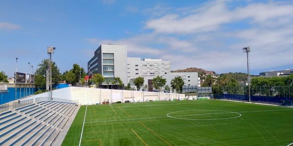 Empty soccer field with bleachers and modern buildings in the background.