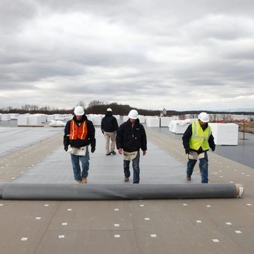 Construction workers walking on a rooftop with a large rolled material.