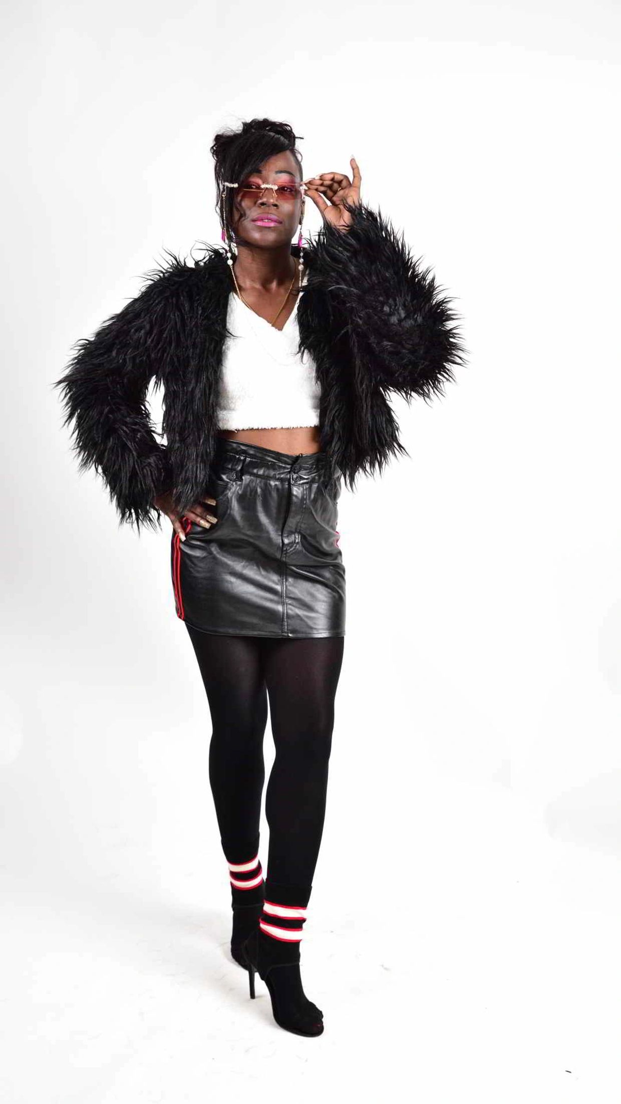 Confident woman in black faux fur jacket and leather skirt adjusting pink sunglasses.