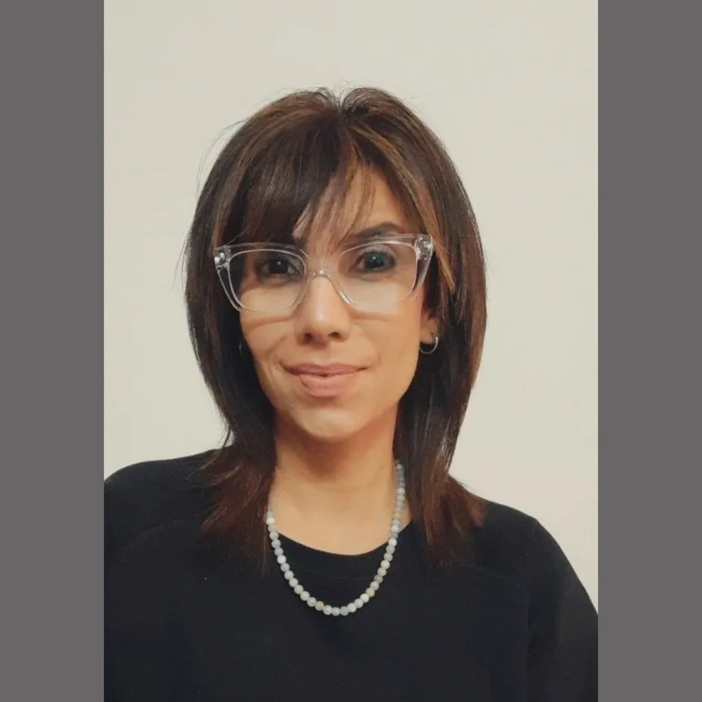 A woman with medium-length brown hair wearing clear glasses and a pearl necklace.