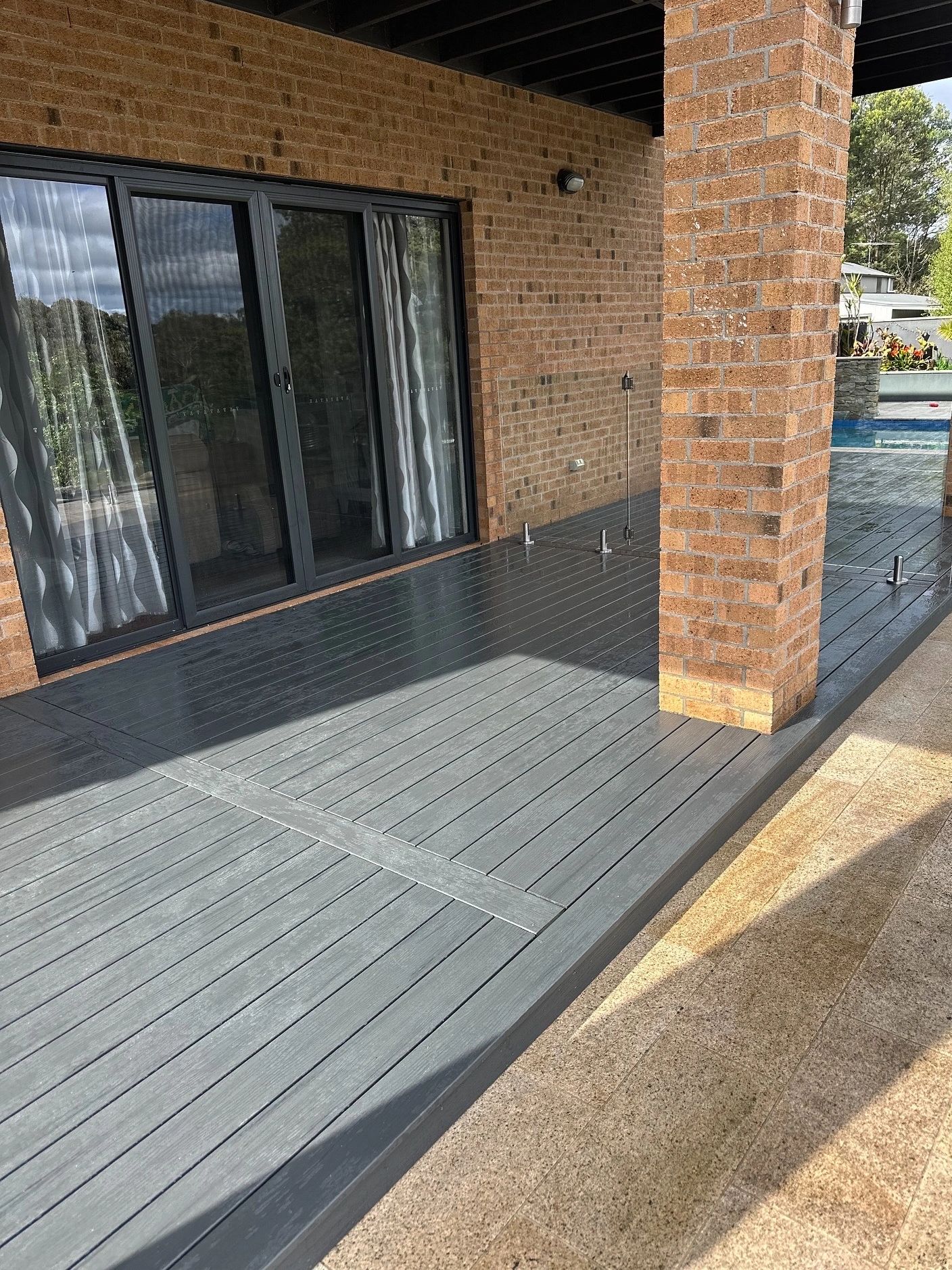 Grey composite deck with a glass balustrade under a covered alfresco beside a brick home