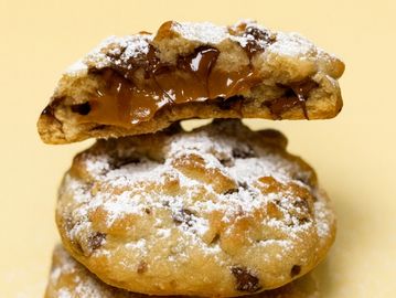 Stack of powdered sugar dusted chocolate chip cookies, one broken showing gooey chocolate.