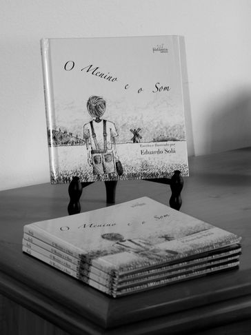 Black and white photo of the book "O Menino e o Som" displayed on a stand and stacked on a table.