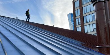 A worker standing on a sloped metal roof with city buildings in the background.