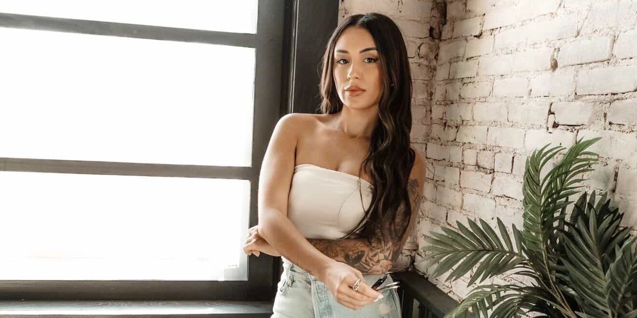 A woman with long hair and tattoos poses confidently near a window and brick wall.