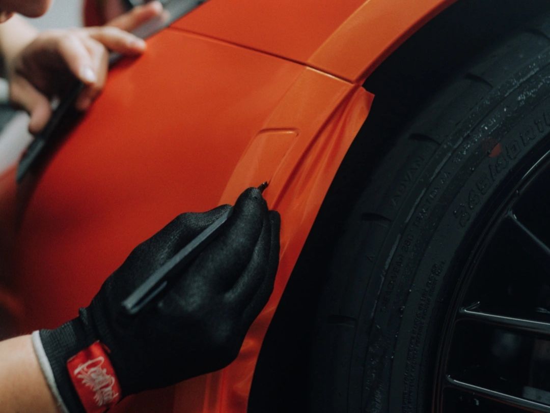 Person applying orange vinyl wrap on a car's fender near the wheel.
