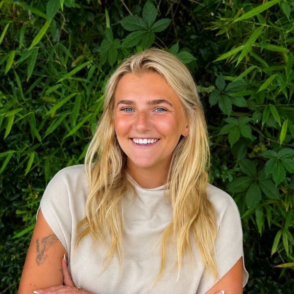 Smiling blonde woman stands confidently in front of lush green foliage.
