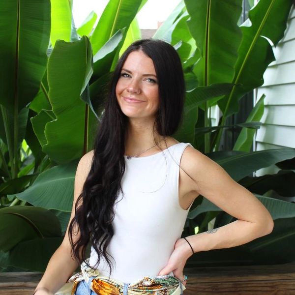 Woman with long dark hair smiling among large green leaves in white top.