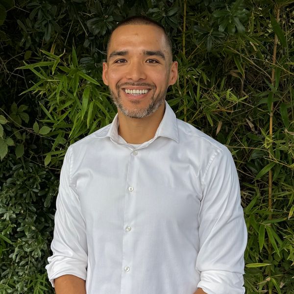 Smiling man with brown hair stands against lush green foliage.